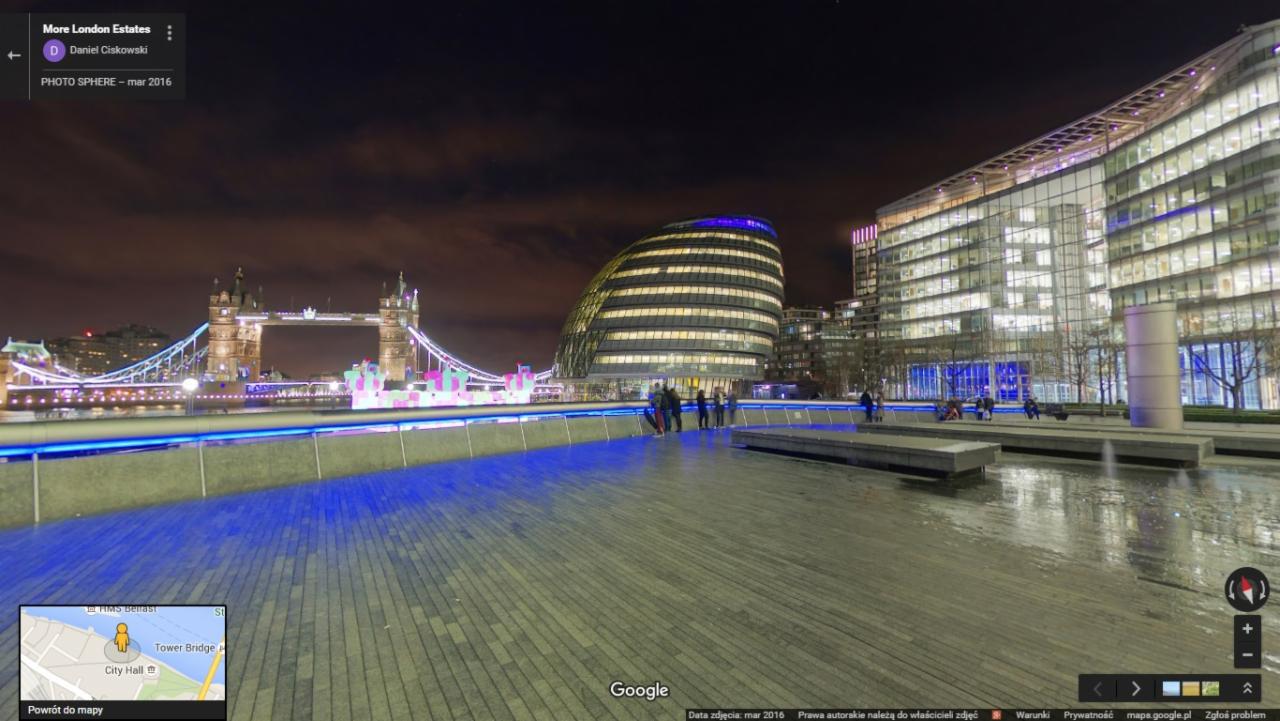 Panoramiczny widok nocny na Tower Bridge i nowoczesne budynki w Londynie, z widoczną iluminacją i odbiciami w mokrej nawierzchni placu.