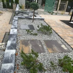 Wystawa materiałów brukowych: ścieżka z kostki w odcieniach beżu, obrzeża z szarej kostki, rabata z grysu i płytami oraz niskimi iglakami, w tle ekspozycja ogrodzeń.