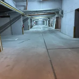 Wnętrze garażu podziemnego z betonową posadzką, widoczne żółto-czarne pasy ostrzegawcze na filarach i instalacje wentylacyjne pod sufitem.