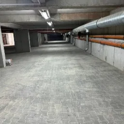 Szara, betonowa kostka brukowa ułożona na parkingu podziemnym, widoczne betonowe filary i rury pod sufitem.