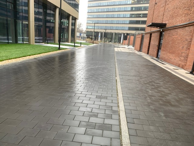 Nowoczesna ścieżka z ciemnej kostki brukowej ułożonej we wzór jodełki, obok trawnik i budynki biurowe z cegły i szkła.