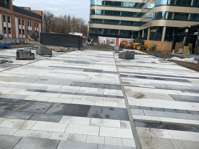 Plac budowy z ułożoną częścią nawierzchni z kostki w odcieniach szarości i bieli, palety z kostką, koparka i budynki w tle.