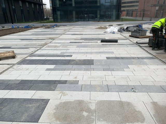 Nowo ułożona nawierzchnia z prostokątnych kostek w odcieniach szarości i bieli, z widocznymi elementami roboczymi i budynkami w tle.