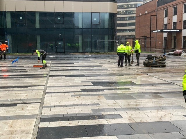 Pracownicy w odblaskowych ubraniach układają wzór z szarych i czarnych kostek na placu przed nowoczesnym budynkiem biurowym w pochmurny dzień. Widoczne narzędzia i paleta z pozostałościami materiału.