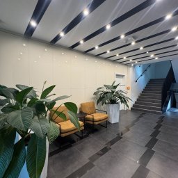 Moppy.pl - Wnętrze biura po sprzątaniu: lśniąca podłoga z płytek, minimalistyczne meble wypoczynkowe, rośliny doniczkowe i nowoczesne oświetlenie sufitowe.