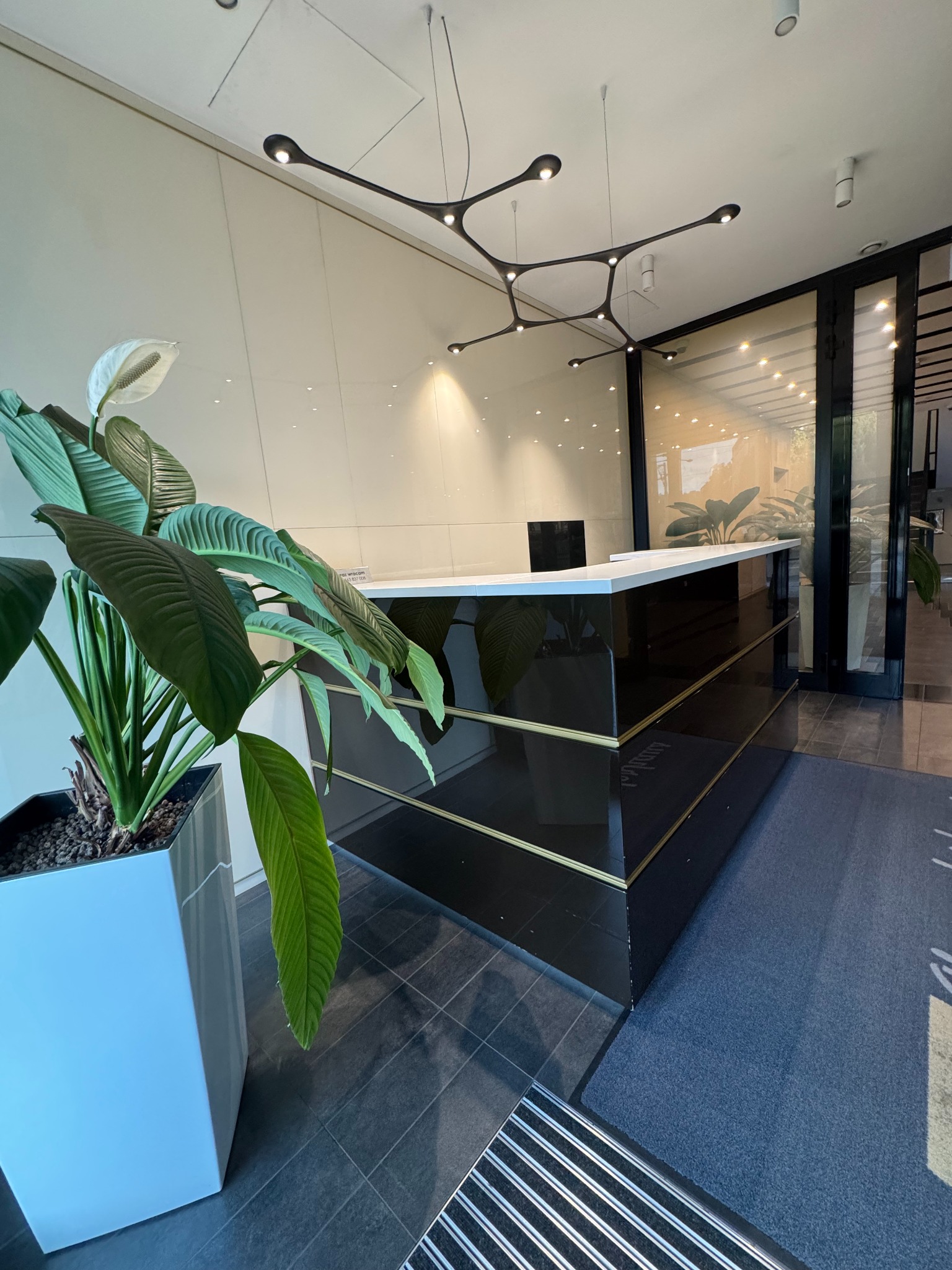 Nowoczesne biuro: czarna lada recepcyjna z białym blatem, designerska lampa sufitowa i roślina w białej donicy. Czyste, jasne wnętrze z elementami szkła.