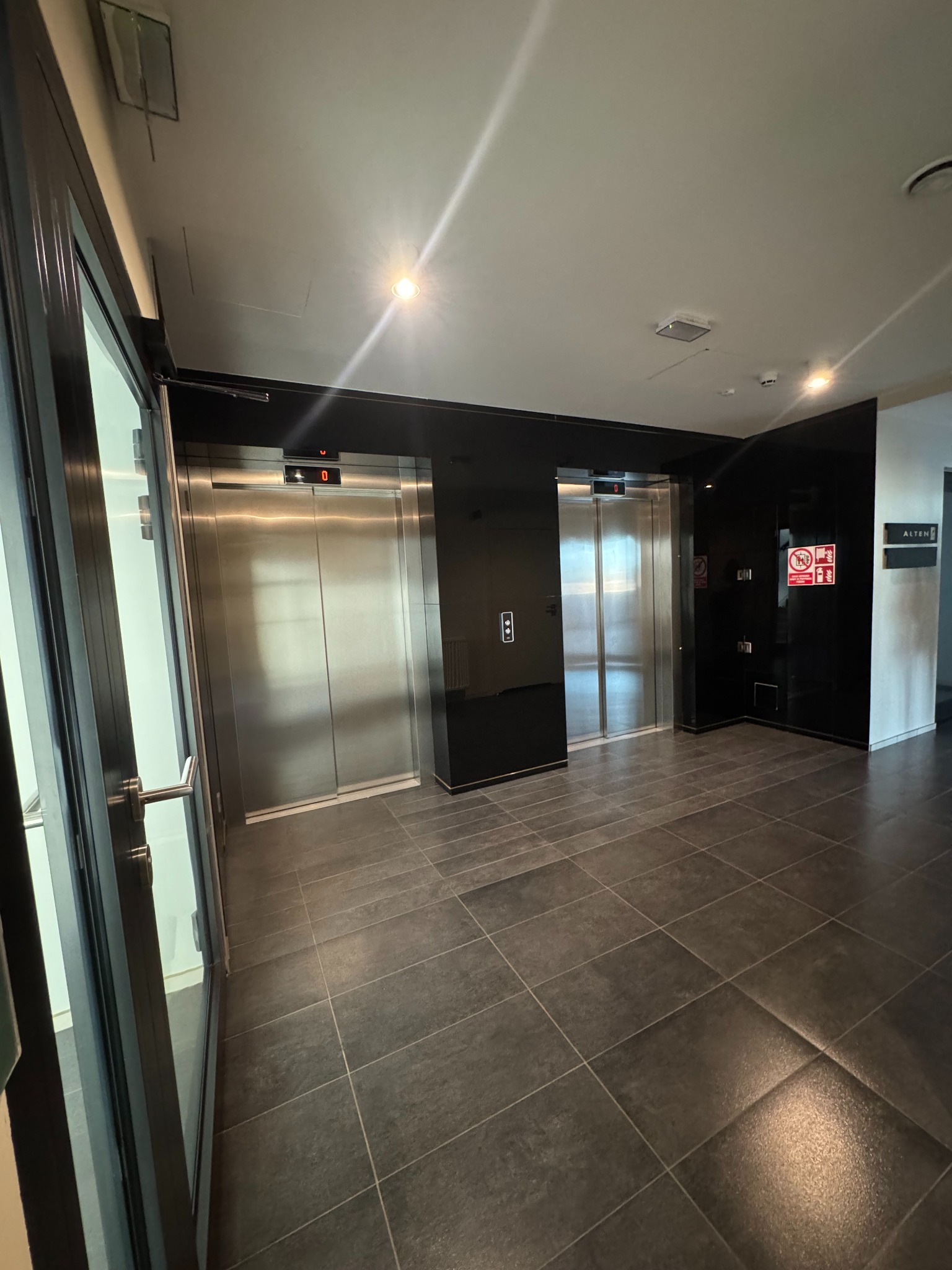 Wnętrze biurowca z windami i ciemną, błyszczącą podłogą z płytek. Widoczne drzwi wejściowe z lewej strony i tabliczka informacyjna na ścianie z prawej.