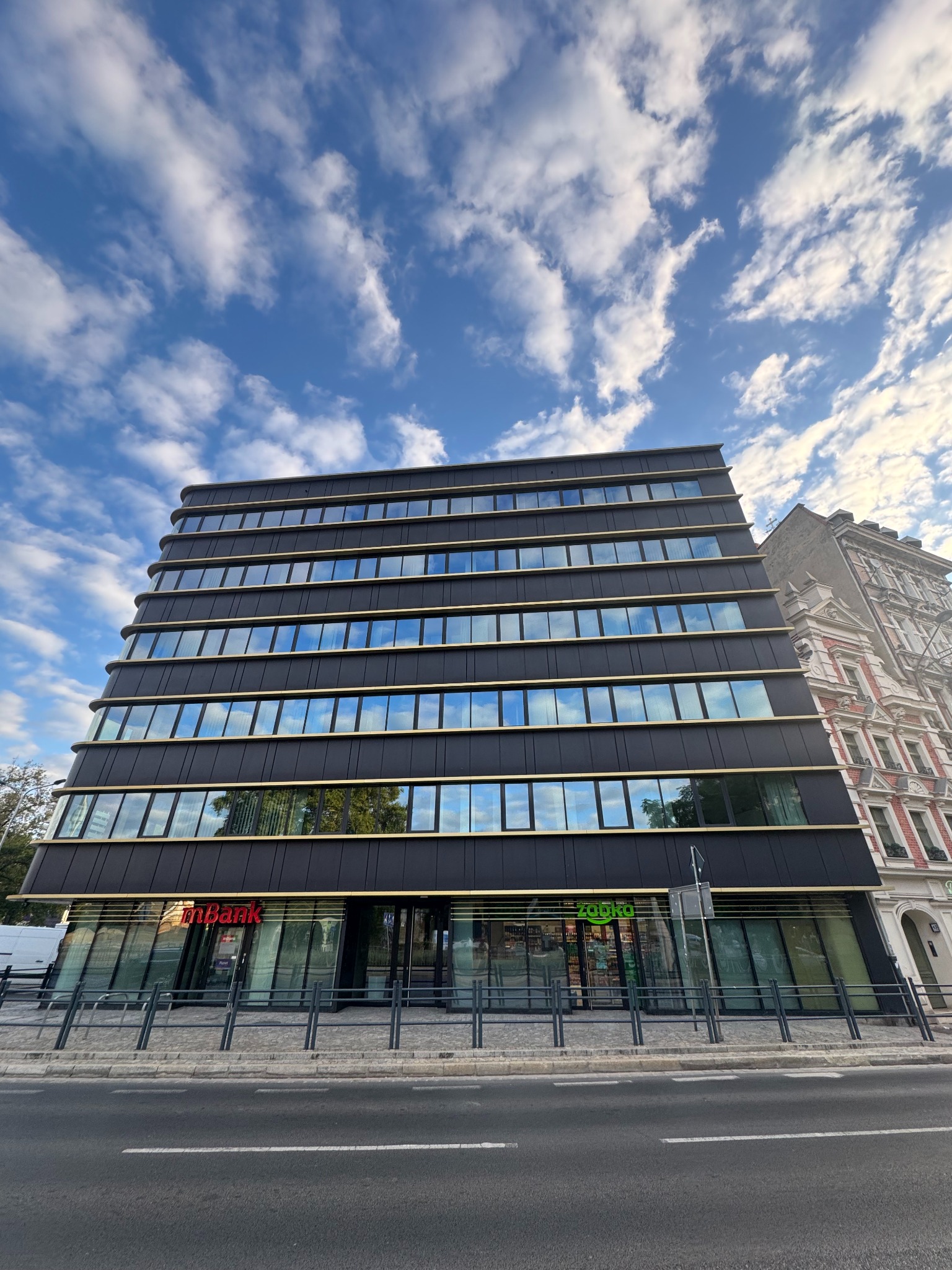 Nowoczesny biurowiec z czarną fasadą i refleksami chmur w oknach, widok z ulicy. Na parterze lokale usługowe z szyldami 'mBank' i 'Żabka'.