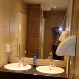 Wykończona łazienka z dwoma umywalkami, czarnym blatem i beżowymi płytkami, widoczny również podajnik na mydło i suszarka do rąk.