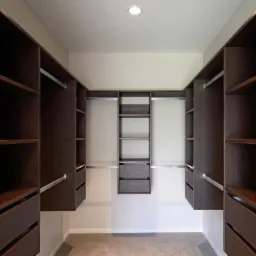 Wnętrze garderoby z zabudową w ciemnym odcieniu drewna, z p&oacute;łkami i szufladami, oświetlone punktowym światłem sufitowym.