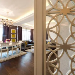 Ażurowy panel dekoracyjny z motywem geometrycznym, oddzielający jadalnię z białym stołem i krzesłami od salonu z szarą sofą, widoczne brązowe zasłony i drewniana podłoga.