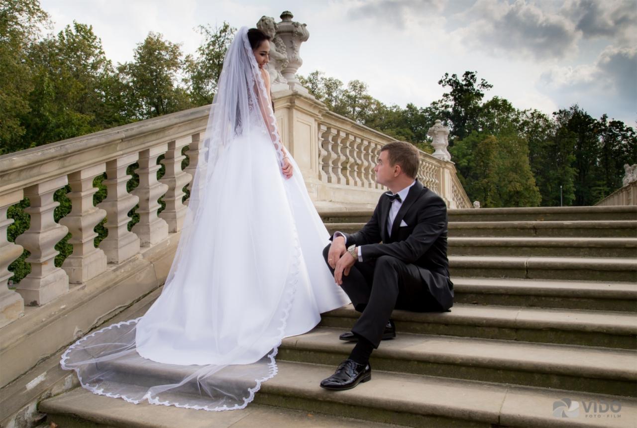 Panna młoda w długiej białej sukni i welonie stoi na schodach, spoglądając na siedzącego obok pana młodego w czarnym garniturze z muchą, na tle balustrady i zieleni.