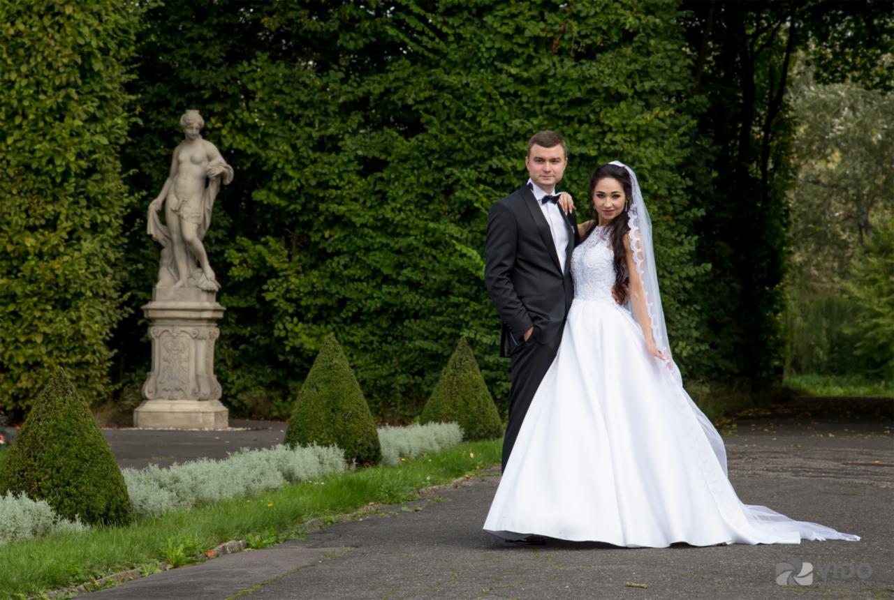 Para młoda pozuje na tle zieleni i klasycznej rzeźby w parku, panna młoda w koronkowej sukni z długim welonem, pan młody w eleganckim smokingu z muchą.