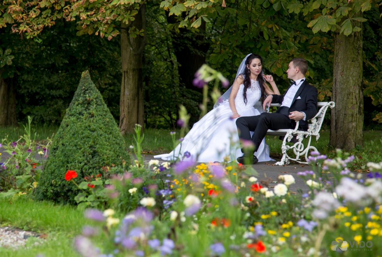 Para młoda siedzi na białej ławce w parku, otoczona kolorowymi kwiatami, z krzewem w kształcie stożka po lewej stronie.