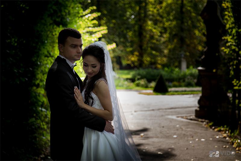 Para młoda w objęciach w zacienionym parku, panna młoda w białej sukni i welonie, pan młody w czarnym smokingu, delikatne światło słoneczne w tle.