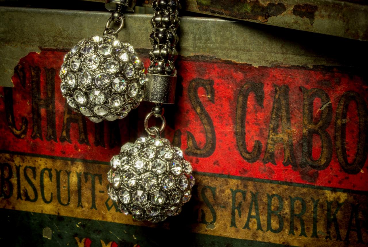 Dwa kolczyki kulki wysadzane cyrkoniami zwisają na tle starej, pomalowanej puszki po ciasteczkach z napisem 'Chas Cabo Biscuit Fabrika'.