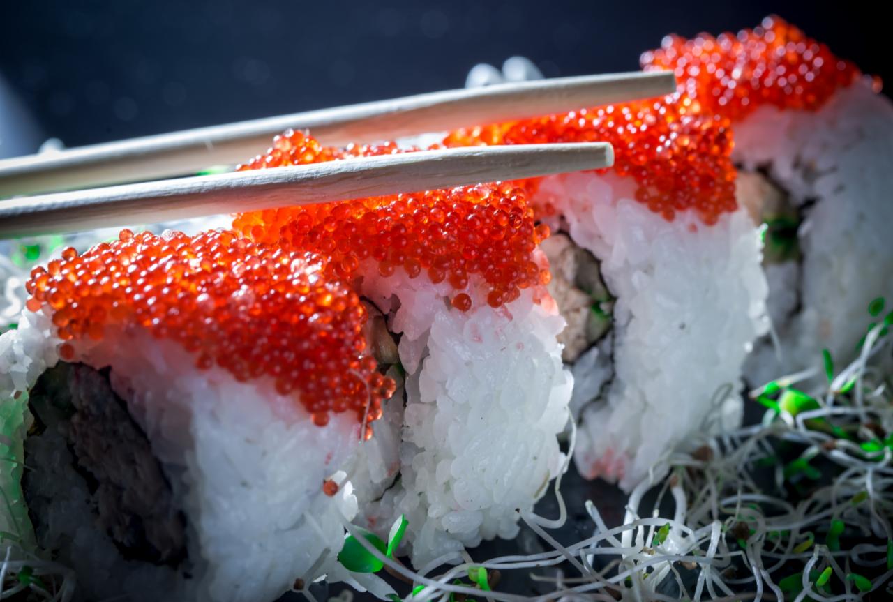 Zbliżenie na rolki sushi z białym ryżem, czerwonym kawiorem i zielonymi kiełkami, podane z pałeczkami na ciemnym tle.