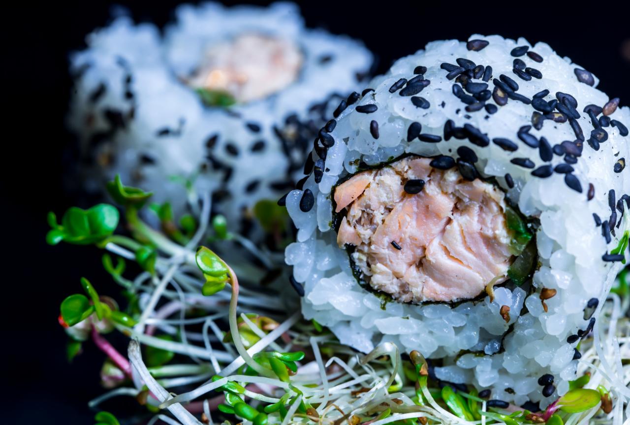 Makro zbliżenie na rolkę sushi z łososiem, ryżem posypanym czarnym sezamem i świeżymi kiełkami na ciemnym tle.
