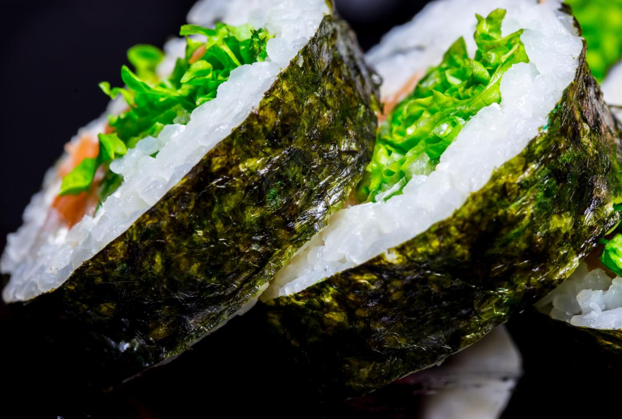 Zbliżenie na dwa kawałki sushi z łososiem i sałatą, owinięte w nori, na czarnym tle.