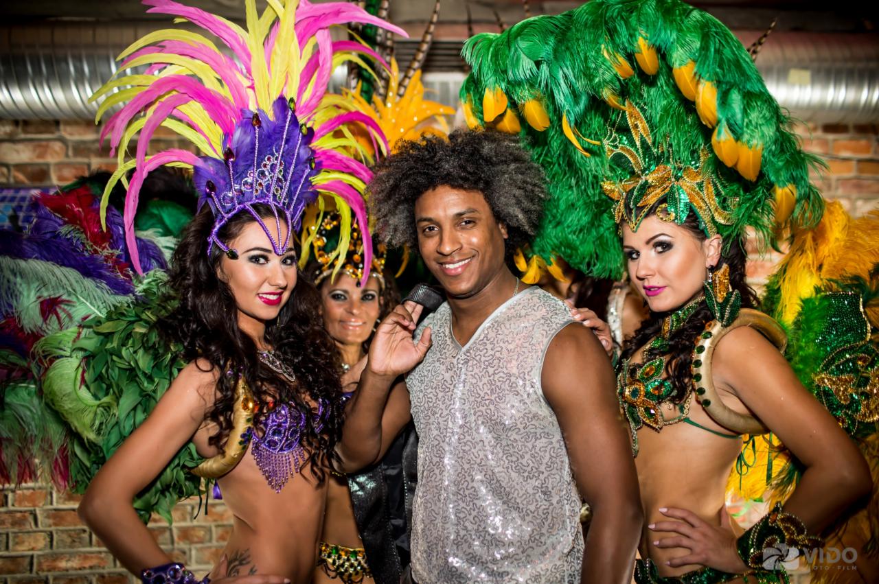 Grupa tancerzy w barwnych, pióropuszowych kostiumach karnawałowych pozuje do zdjęcia, z uśmiechem prezentując się na tle ceglanej ściany.