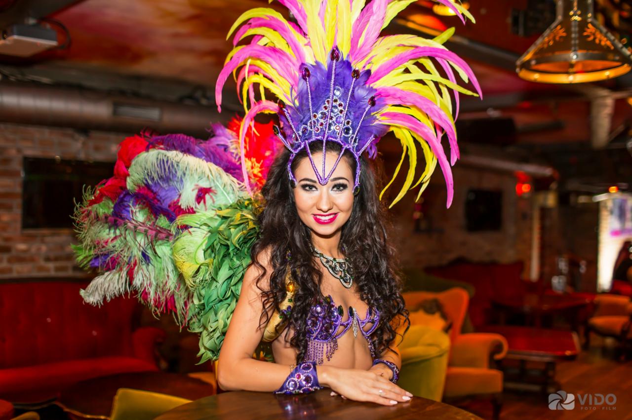 Uśmiechnięta kobieta w bogatym stroju karnawałowym z piórami, cekinami i kryształkami, oparta o okrągły stolik w klubowym wnętrzu z czerwonymi sofami i ceglaną ścianą.