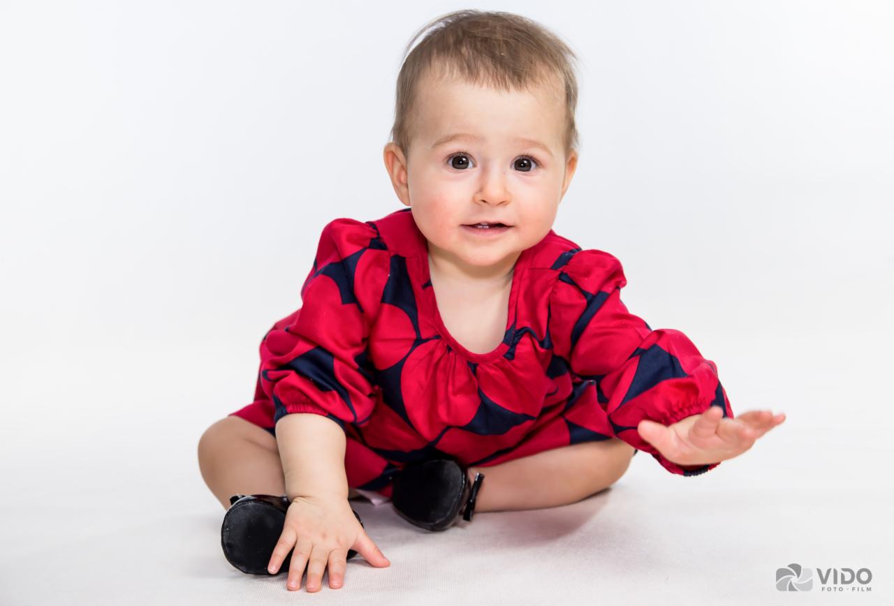 Uśmiechnięta, siedząca dziewczynka w czerwonej sukience z granatowymi grochami, na białym tle studyjnym.