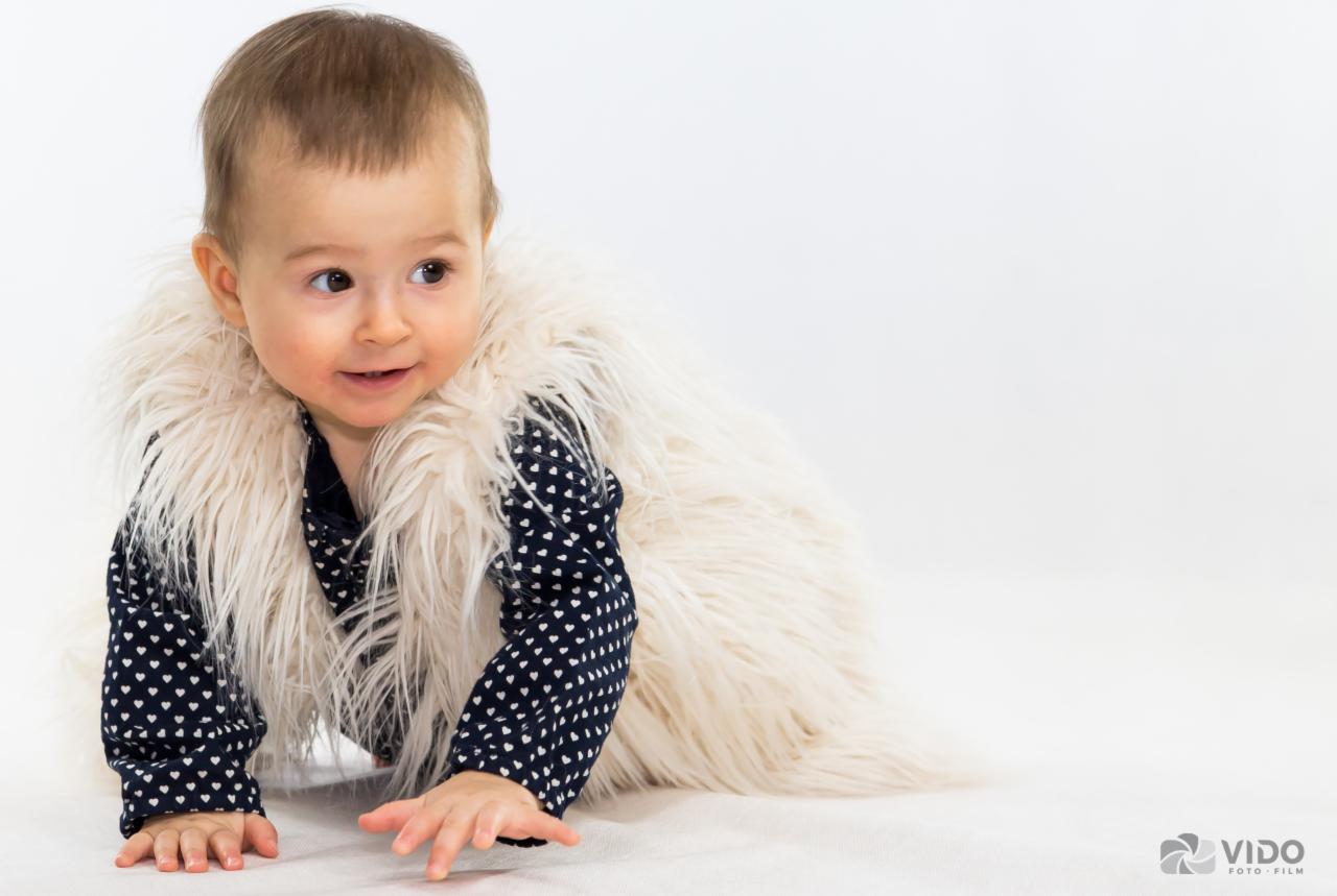 Uśmiechnięty niemowlak w ciemnej bluzce w białe serduszka i futrzanej kamizelce na białym tle, raczkuje w stronę widza.