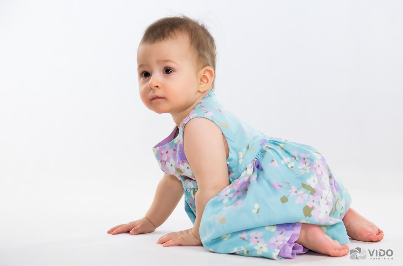 Pełzające dziecko w błękitnej sukience w kwiaty na białym tle, spoglądające w lewo.