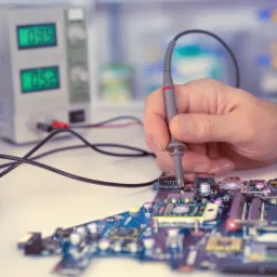 Miernik w dłoni przyłożony do niebieskiej płyty głównej komputera na tle laboratoryjnego zasilacza z wyświetlaczami LED.