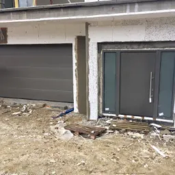 Szara brama garażowa segmentowa i szare drzwi wejściowe z bocznymi panelami szklanymi w trakcie montażu, widoczne narzędzia i materiały budowlane na placu budowy.