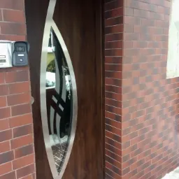 Zewnętrzne drzwi wejściowe w Pruszczu Gdańskim, z brązowego drewna z pionowym przeszkleniem w kształcie łzy, obramowane metalową listwą. Drzwi zamontowane w murze z czerwonej cegły.