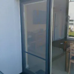 Szara przesuwna moskitiera w ramie zamontowana na drzwiach balkonowych, widok z zewnątrz na taras z krzesłem i czarną wycieraczką.