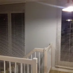 Dwa okna z białymi żaluzjami poziomymi, widoczne zza białej drewnianej balustrady schodów, oświetlone górnym światłem.