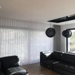 Nowoczesny salon z dużymi oknami przesłoniętymi długimi, białymi, transparentnymi zasłonami, dopełniony czarną skórzaną sofą i trzema okrągłymi lampami wiszącymi o ażurowej strukturze.