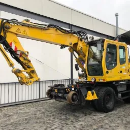 Żółta koparko-ładowarka kołowa z dodatkowym osprzętem kolejowym, zaparkowana na zewnątrz budynku z szarą bramą i białymi panelami ściennymi.