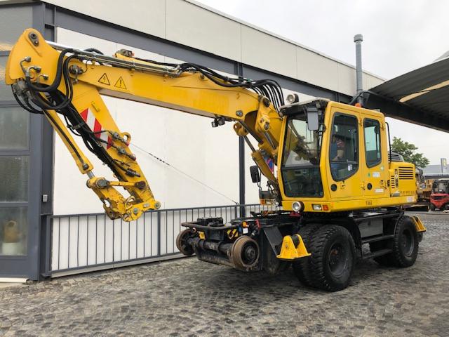 Żółta koparko-ładowarka kołowa z dodatkowym osprzętem kolejowym, zaparkowana na zewnątrz budynku z szarą bramą i białymi panelami ściennymi.