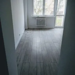 Pusty pokój po remoncie z nową podłogą z paneli imitujących drewno, białymi ścianami i grzejnikiem pod oknem z widokiem na drzewa.