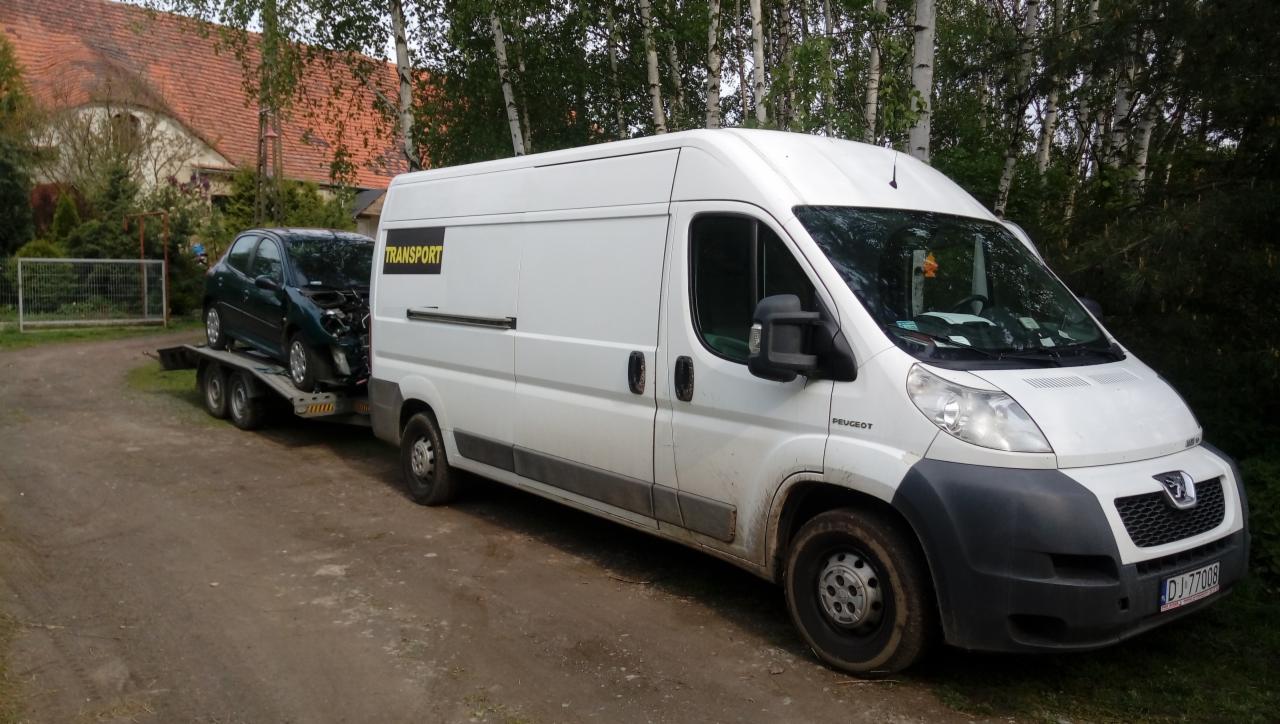 Biały bus Peugeot z przyczepą, na której znajduje się uszkodzony zielony samochód osobowy, zaparkowany na żwirowej drodze przed budynkiem.