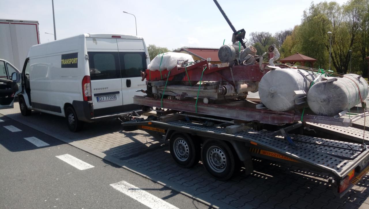 Biały bus Peugeot z napisem 'Transport' zaparkowany obok przyczepy z załadowaną maszyną przemysłową zabezpieczoną pasami, widok z boku na zewnątrz.