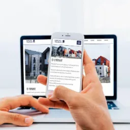 Prezentacja strony internetowej firmy budowlanej na laptopie i smartfonie, ukazująca nowoczesne budynki mieszkalne w tle, z widoczną filiżanką kawy po prawej stronie.