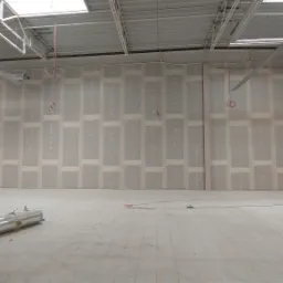 Wnętrze remontowanego, dużego pomieszczenia, prawdopodobnie sklepu. Ściany przygotowane pod dalsze prace, z widocznymi pasami szpachli i płyt gipsowo-kartonowych. Na podłodze leżą rury wentylacyjne...