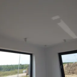 Wnętrze nowo wykończonego pokoju z białymi ścianami i sufitem, widoczne okna z czarnymi ramami i punktowe oświetlenie sufitowe.