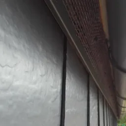 Szczegółowy widok na dach pokryty ciemnymi płytami łupkowymi, z widoczną metalową siatką wentylacyjną okapu i fragmentem rynny z PCV, uchwycony pod kątem z dołu.