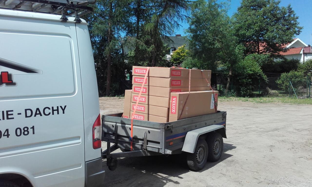 Przyczepka samochodowa wypełniona kartonami okien dachowych marki Velux, ciągnięta przez biały samochód dostawczy z napisem '...KIE - DACHY' i numerem telefonu, na tle zieleni drzew i domów.