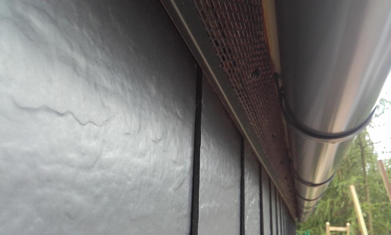 Szczegółowy widok na dach pokryty ciemnymi płytami łupkowymi, z widoczną metalową siatką wentylacyjną okapu i fragmentem rynny z PCV, uchwycony pod kątem z dołu.
