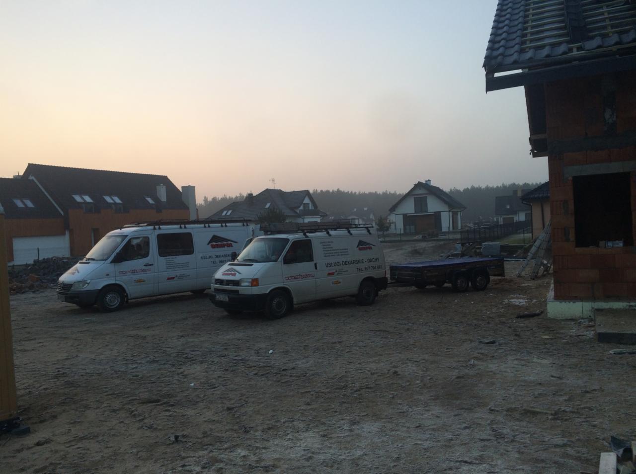 Dwa białe busy z logo firmy dekarskiej i przyczepą na placu budowy z domami w tle, widok wczesnym rankiem.