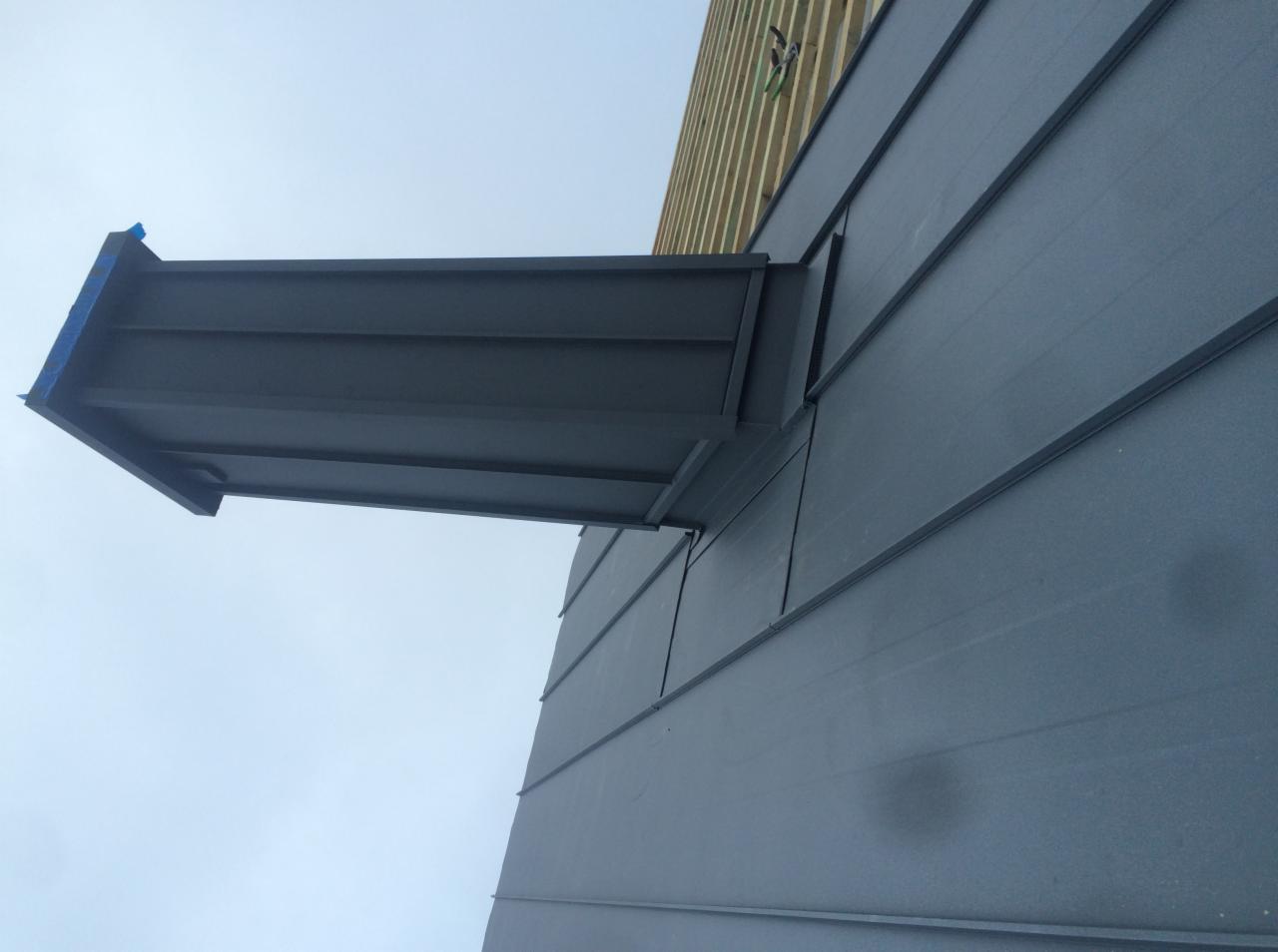 Szary dach z blachy na rąbek stojący z zamontowanym kominem wentylacyjnym, widok z dołu na fragment dachu i niebo.