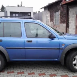 Błękitny Land Rover Freelander zaparkowany na brukowanym podjeździe przed budynkiem z czerwonymi drzwiami garażowymi. Widoczny dach i okna sąsiednich budynków.