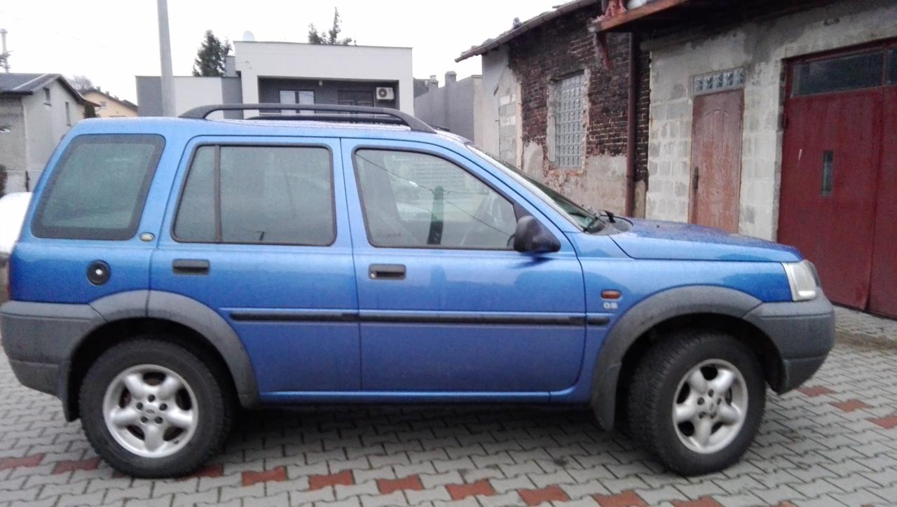 Błękitny Land Rover Freelander zaparkowany na brukowanym podjeździe przed budynkiem z czerwonymi drzwiami garażowymi. Widoczny dach i okna sąsiednich budynków.