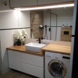 Nowoczesna łazienka z białą szafką pod umywalkę z drewnianym blatem, na którym stoi biała umywalka nablatowa i dekoracyjna orchidea, obok pralka, w tle kabina prysznicowa z czarnymi ramami...